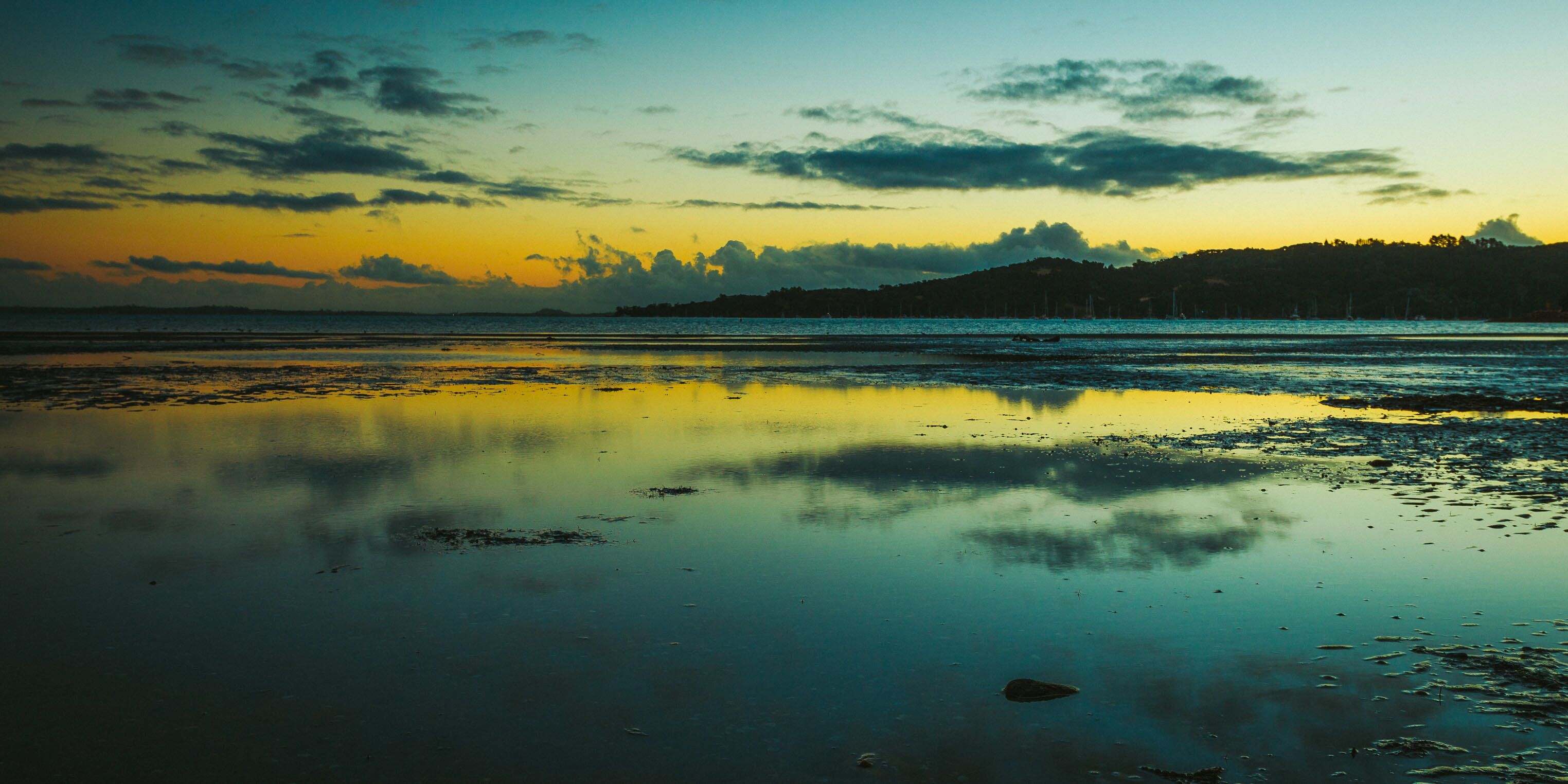 Una playa con aguas tranquilas que reflejan el cielo, una costa lejana con árboles y un horizonte despejado.