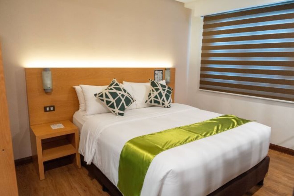 A hotel room with a wooden headboard, a bedside table, a bed with white bedding and a green throw, and a window with horizontal blinds.