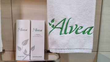 Un baño con un perchero metálico que sostiene una toalla blanca con el bordado 'Alvea' en verde, y dos cajas blancas con el nombre 'Alvea' y un símbolo de kit de bienvenida.
