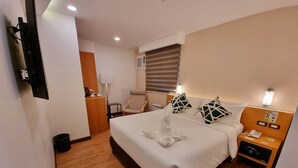 A hotel room with a large bed, a bedside table with a phone and a lamp, a chair, a desk with a coffee maker, and a trash bin. - Alvea Hotel (Puerto Princesa)