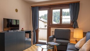 Un soggiorno moderno con un divano, un tavolino in vetro, una TV su un mobile e vista su una foresta attraverso ampie finestre.