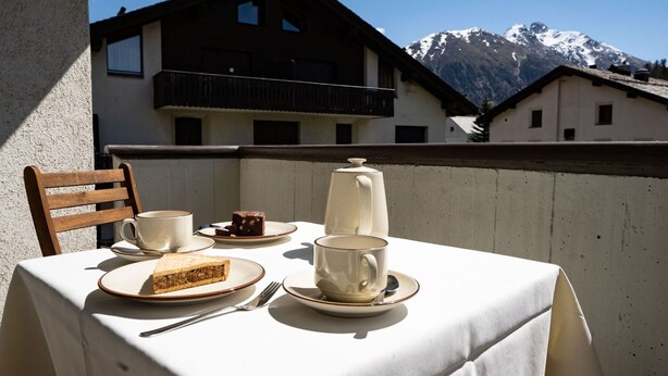 Un tavolo apparecchiato con una fetta di torta, una tazza e un teiera su un balcone con vista sulle montagne.
