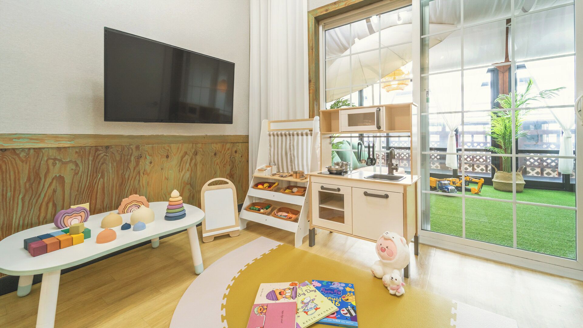 A child's playroom with a play table, toys, a small kitchen set, and a large window with a view of a garden.