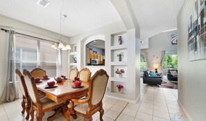 A dining area with a wooden table and chairs, a kitchen in the background, and a living area with a sofa and lamp.