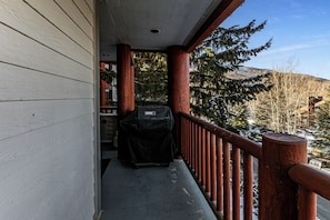 Une véranda couverte avec un barbecue, une balustrade en bois et une vue sur des arbres et des collines enneigés.