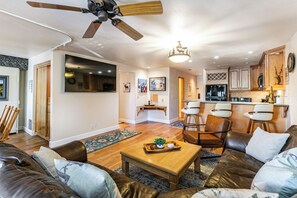 A modern living room with a ceiling fan, a dining area with bar stools, and a kitchen with wooden cabinets and a large refrigerator.