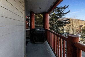 Une véranda couverte avec un barbecue, une balustrade en bois et une vue sur des arbres et des collines enneigés.