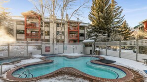 Una piscina con borde de ladrillo, rodeada de nieve y contigua a un edificio con balcones.