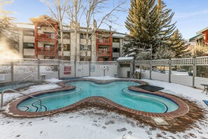 Una piscina con bordo in mattoni, circondata dalla neve, adiacente a un edificio con balconi.