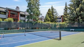 Una cancha de tenis al aire libre con una red, rodeada por una cerca verde y edificios residenciales al fondo.