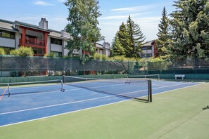 Un campo da tennis all'aperto con una rete, circondato da una recinzione verde, e edifici residenziali sullo sfondo.