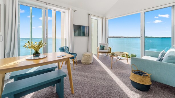 A modern living room with a large window overlooking a body of water, a wooden table, blue chairs, a sofa, and a TV mounted on the wall.