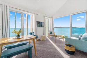 Un salon moderne doté d’une grande fenêtre donnant sur un plan d’eau, d’une table en bois, de chaises bleues, d’un canapé et d’un téléviseur fixé au mur.