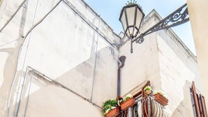 A narrow street with a building featuring a small balcony with potted plants, a door with a number, and a street lamp. - Holiday Home Sleeps 6 in the Historic Center of Le (Lecce)