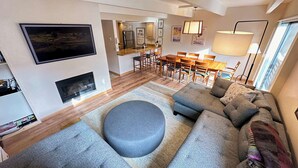 A modern living room with a grey sectional sofa, a round ottoman, a flat-screen TV mounted on the wall, a fireplace, and a dining area with wooden furniture. - Vail Mountain Views From Upper Level 2BR Condo - In Unit Washer/Dryer (Vail)