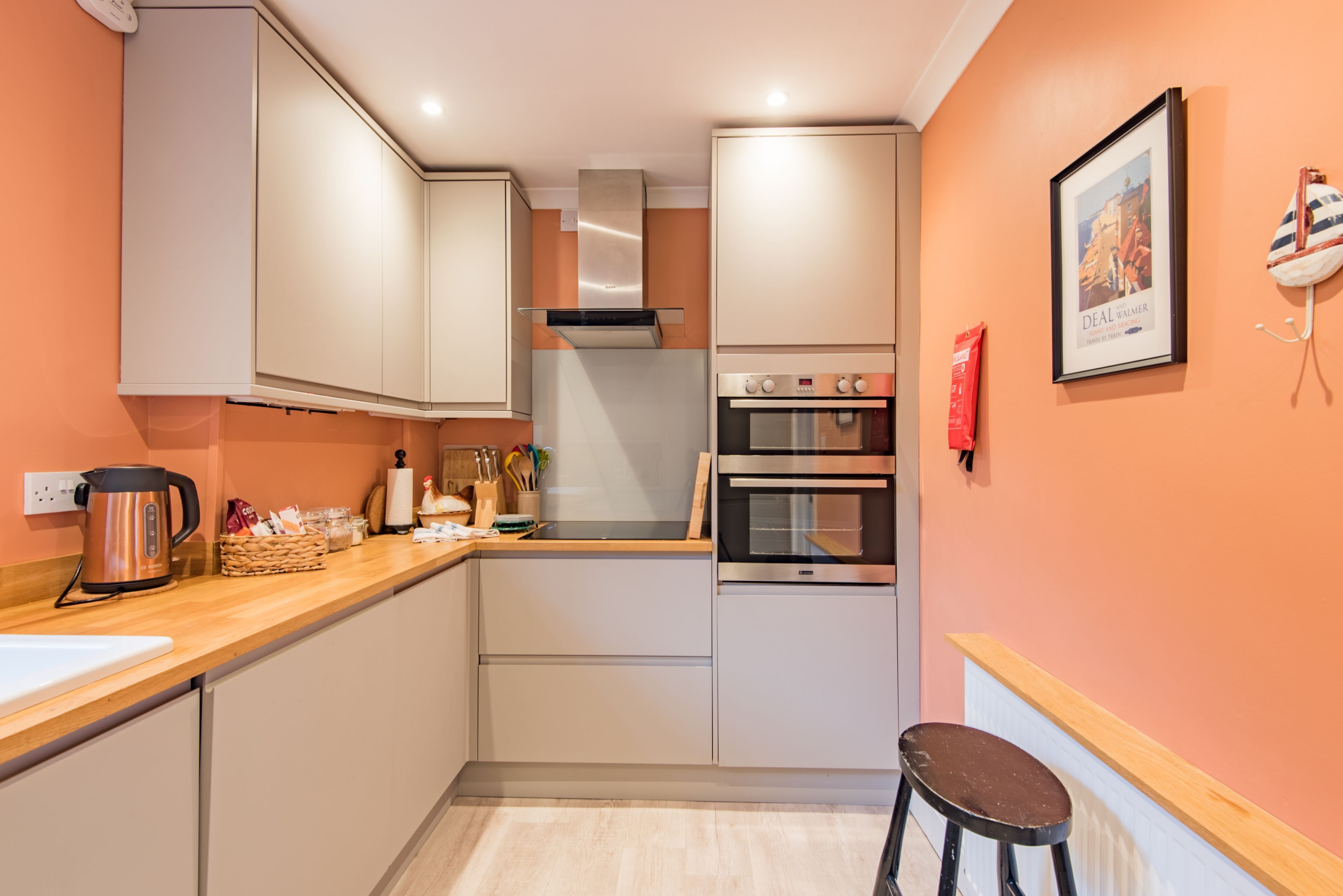 Una cocina compacta con armarios blancos, una campana extractora de acero inoxidable, un horno doble y un taburete pequeño.