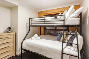 A bunk bed with a wooden headboard, a bedside table with a lamp, and a window with blinds.