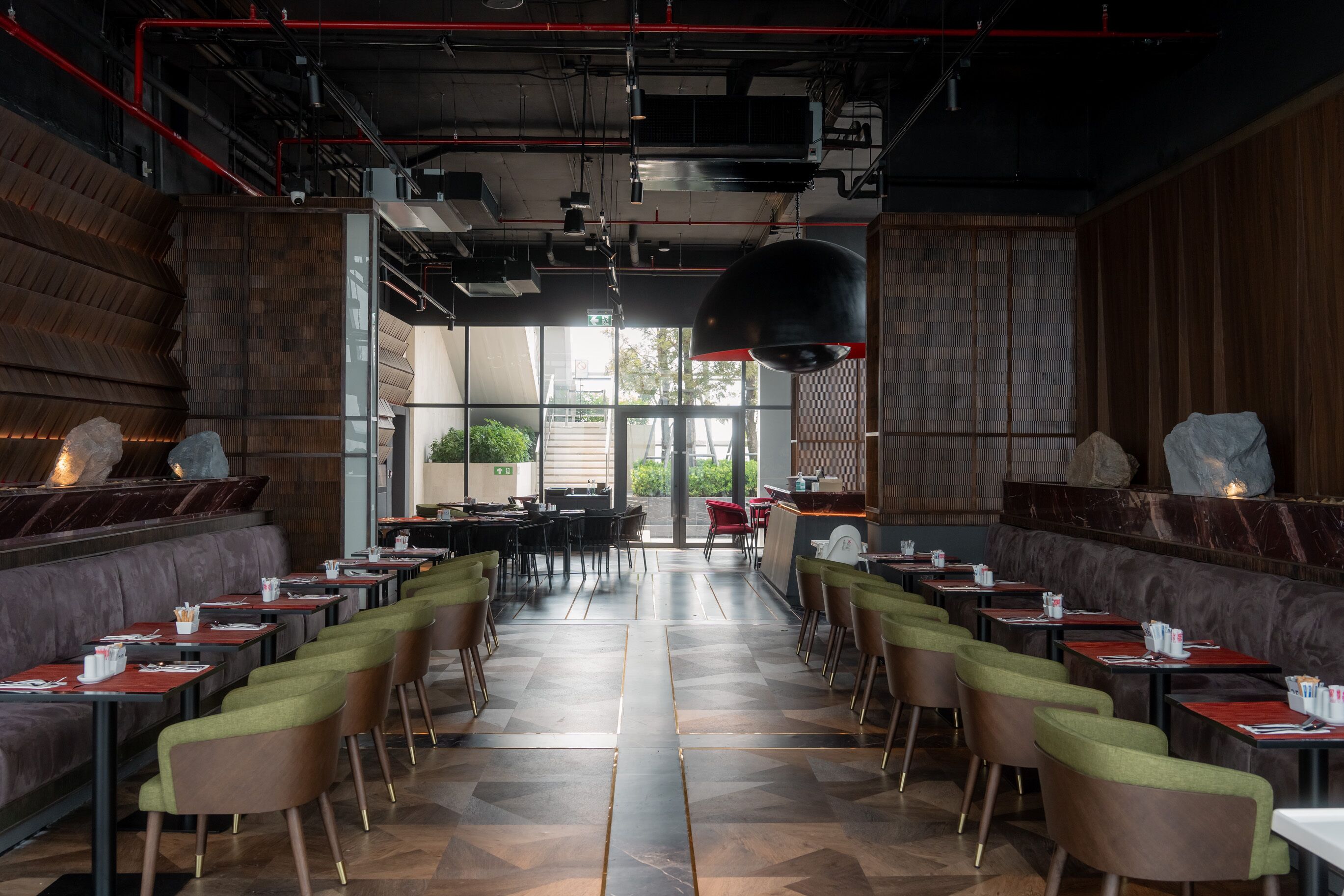 A restaurant with a modern interior, featuring a long counter with booths, a high ceiling with exposed pipes, and a view of the outdoors through large windows.