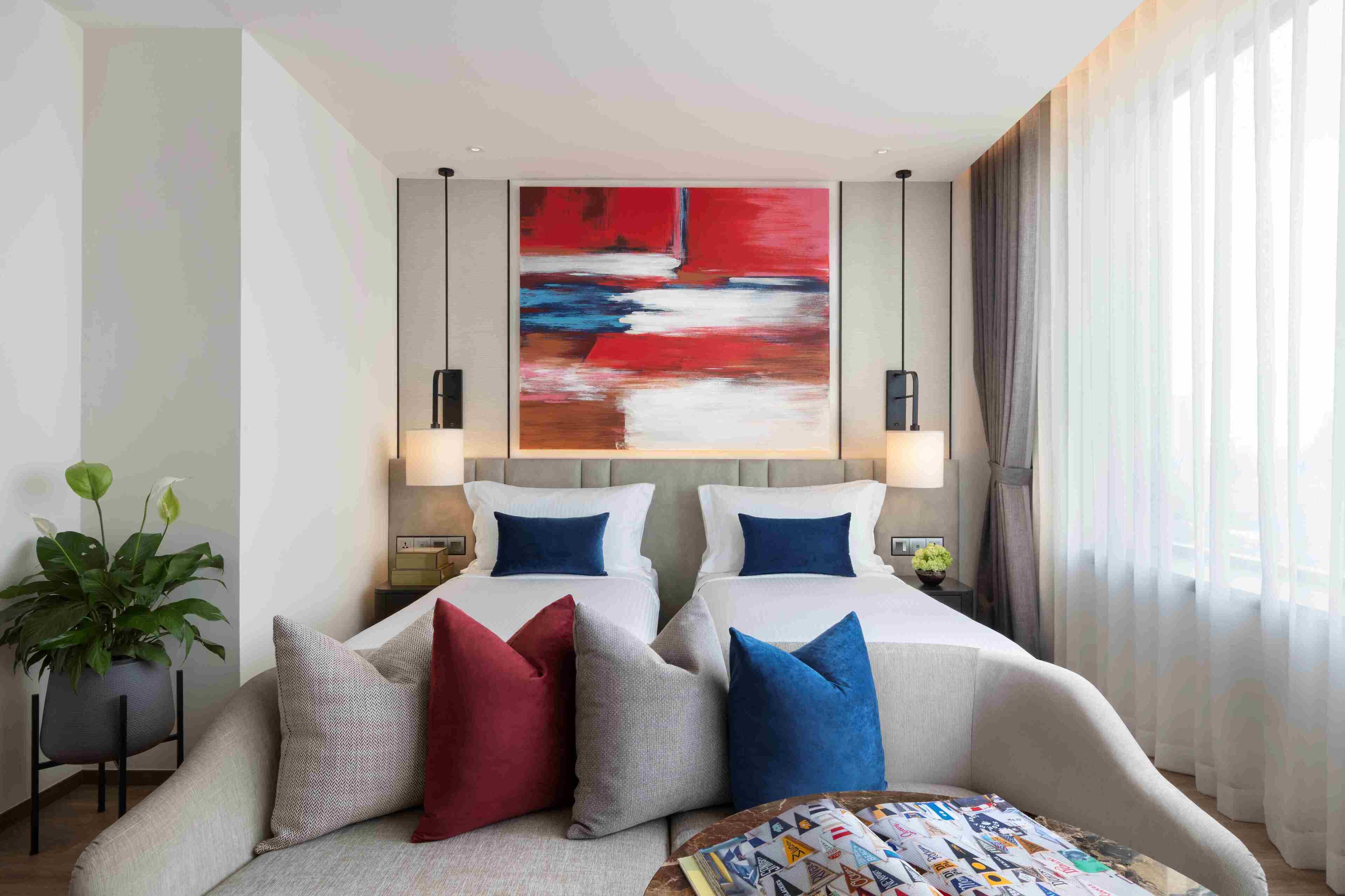 Une chambre d’hôtel avec un grand lit, deux tables de chevet, un canapé, un grand tableau abstrait et une plante en pot.