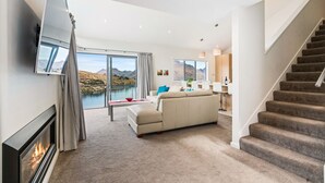 A modern living room with a sofa, a flat-screen TV, a fireplace, and a view of a lake and mountains. - Lakeview Retreat - Queenstown Holiday Home (Queenstown)