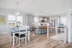 A modern kitchen with white cabinetry, a central island with bar stools, and a dining area with a white table and chairs. - Private Saltwater Pool (Heating Optional), With Excellent Ocean Views Only 2 Blocks From the Beach (Oak Island)