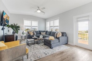 A spacious living room with a grey sectional sofa, a coffee table, and a yellow armchair. There is a ceiling fan, large windows with blinds, and a view of the outdoors. - Private Saltwater Pool (Heating Optional), With Excellent Ocean Views Only 2 Blocks From the Beach (Oak Island)