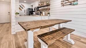 Una cocina con una mesa y banco de madera, muebles blancos y un estante en la pared con objetos decorativos.