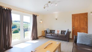 A living room with a sofa, a wooden table, and a view through a large window. - Croftdean -  a house that sleeps 6 guests  in 3 bedrooms (Dalbeattie)