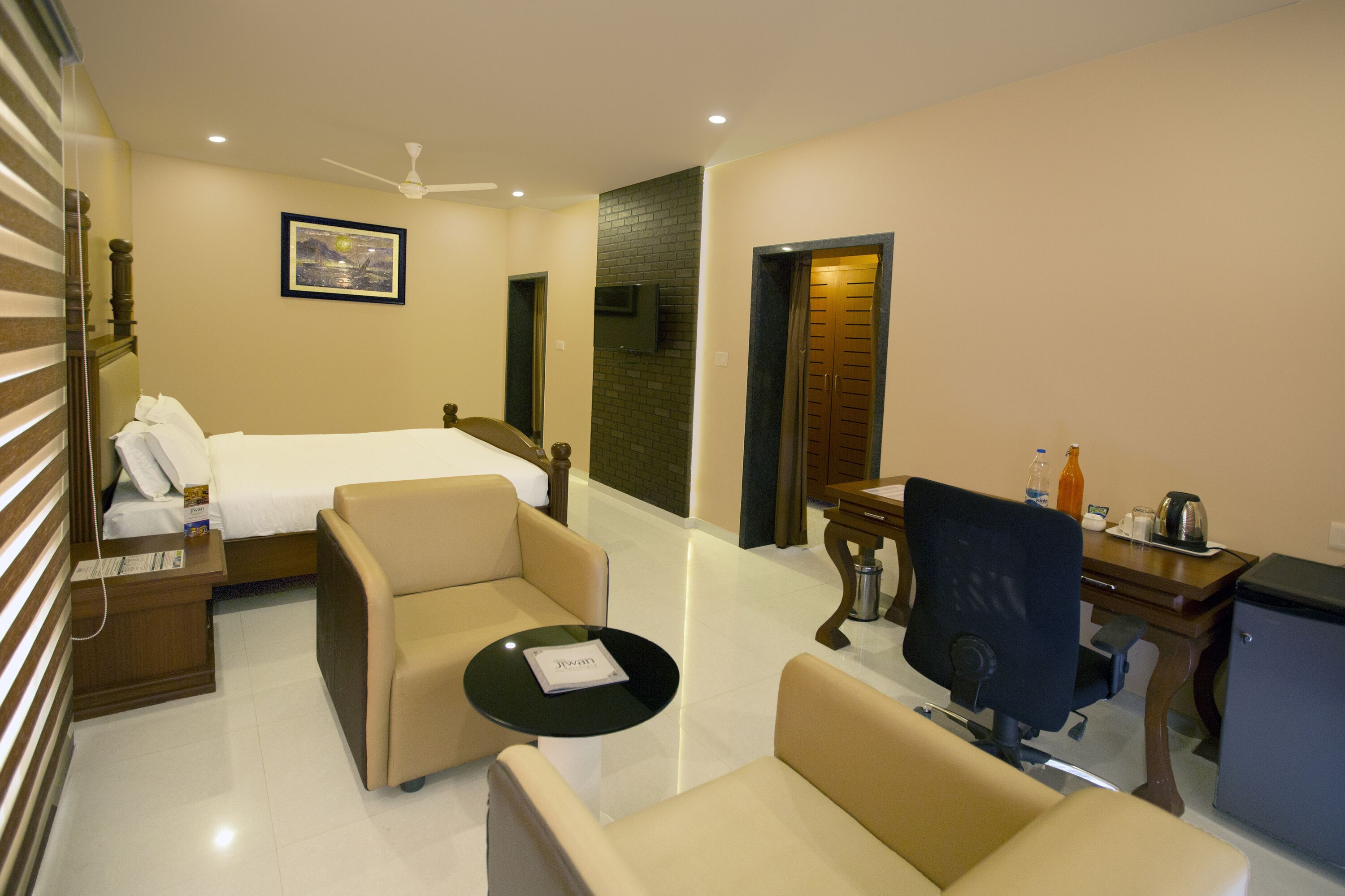 Habitación de hotel con cama, dos sillones, una mesita, escritorio con computadora, cafetera y televisor.