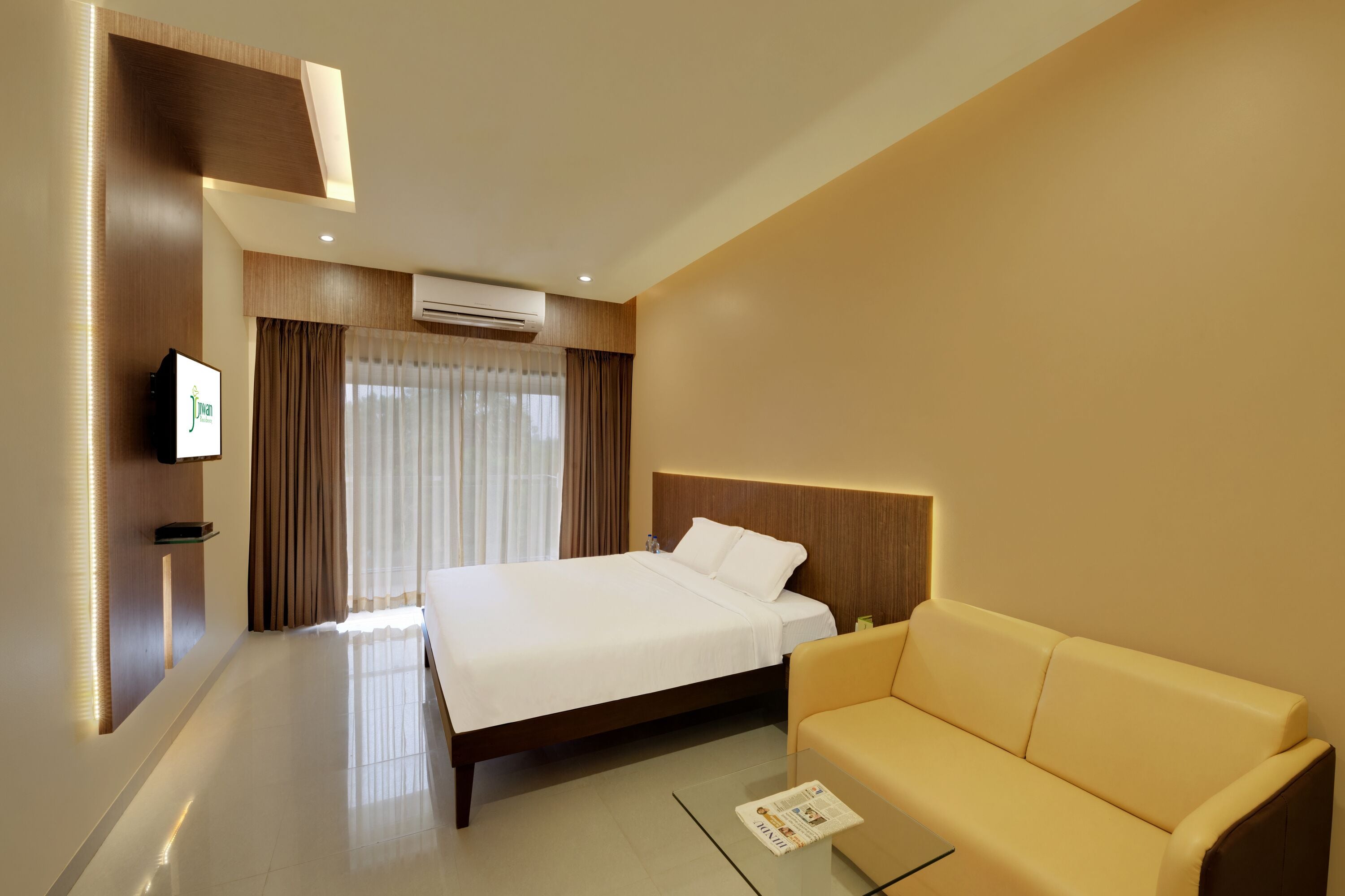 A hotel room with a bed, a sofa, a flat-screen TV, and an air conditioning unit.