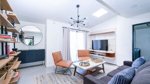 Een moderne woonkamer met een bank, stoelen, een salontafel en een televisie. Er staan boekenkasten met boeken en er hangt een spiegel.