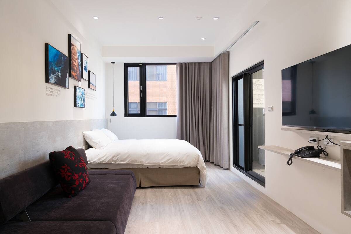 A modern hotel room with a bed, a sofa, a TV, and artwork on the wall.