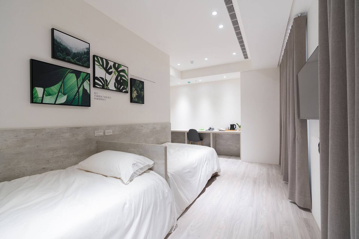 A modern hotel room with two beds, a desk, and a view of the city.