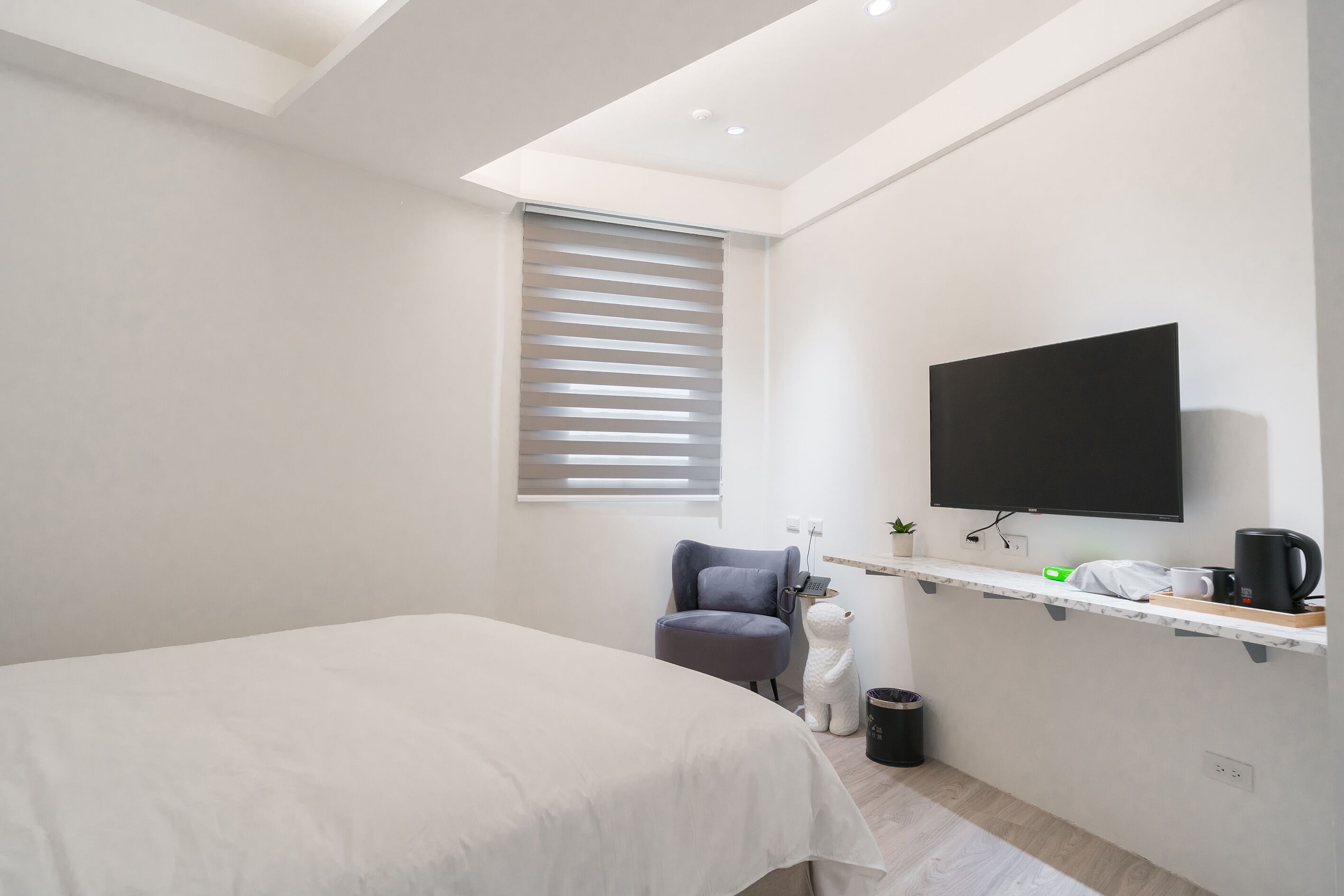 A modern hotel room with a bed, a chair, a TV, a small shelf with a coffee maker, and a trash bin.