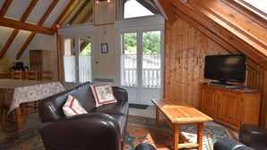 Un acogedor cuarto ático con techo de madera, televisor, sofá, mesa de comedor y vistas al exterior a través de amplias ventanas.