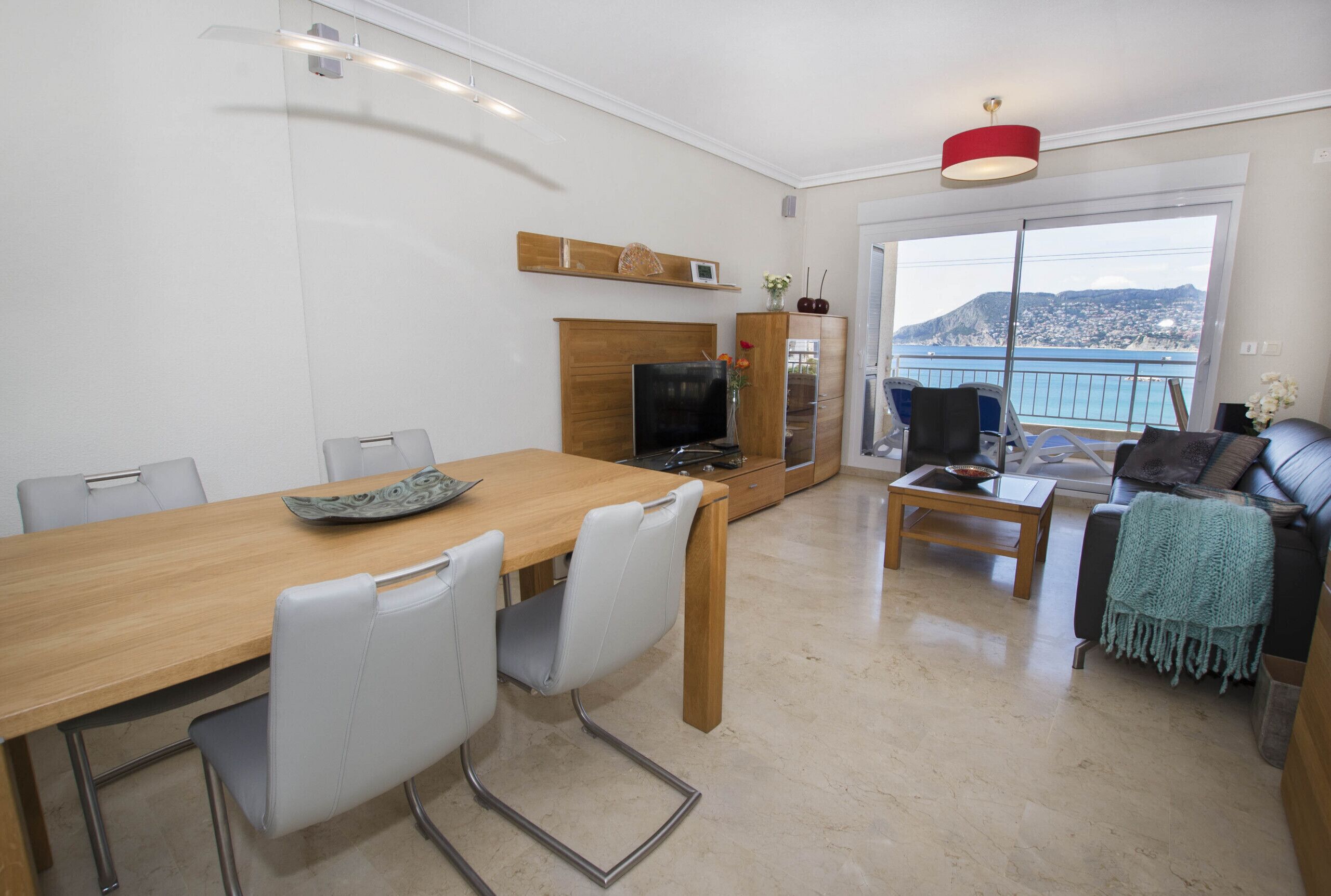 Uma sala de estar moderna com uma mesa de jantar de madeira, cadeiras brancas, um sofá e uma TV. Há uma janela grande com vista para o mar.