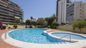 Une piscine avec une terrasse carrelée, entourée de palmiers et bordée d’un immeuble à plusieurs étages doté de balcons.