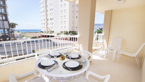Un balcon avec une table ronde pour quatre personnes, des chaises blanches et une vue sur la mer et les bâtiments.