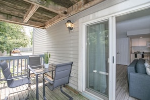 Relax on the covered patio with a glass table and two chairs, surrounded by a residential interior with a sofa and kitchen area.