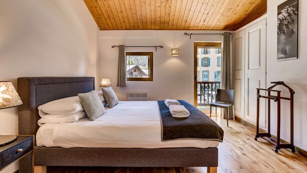 Ein Schlafzimmer mit Holzboden, einem Bett mit weißen Leinentüchern, einem Nachttisch mit Lampe, einem Fenster mit Vorhängen und Blick auf Gebäude im Freien.
