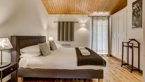 Ein Schlafzimmer mit Holzbalkendecke, einem Bett mit dunklem Kopfteil, zwei Nachttischlampen, einer Wandleuchte und einem Fenster mit Vorhängen.
