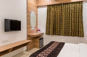 A hotel room with a bed, a wooden desk, a television, a mirror, a small sink, and a window with curtains. - Meridian hotel Banquets and Rooms (Surat)