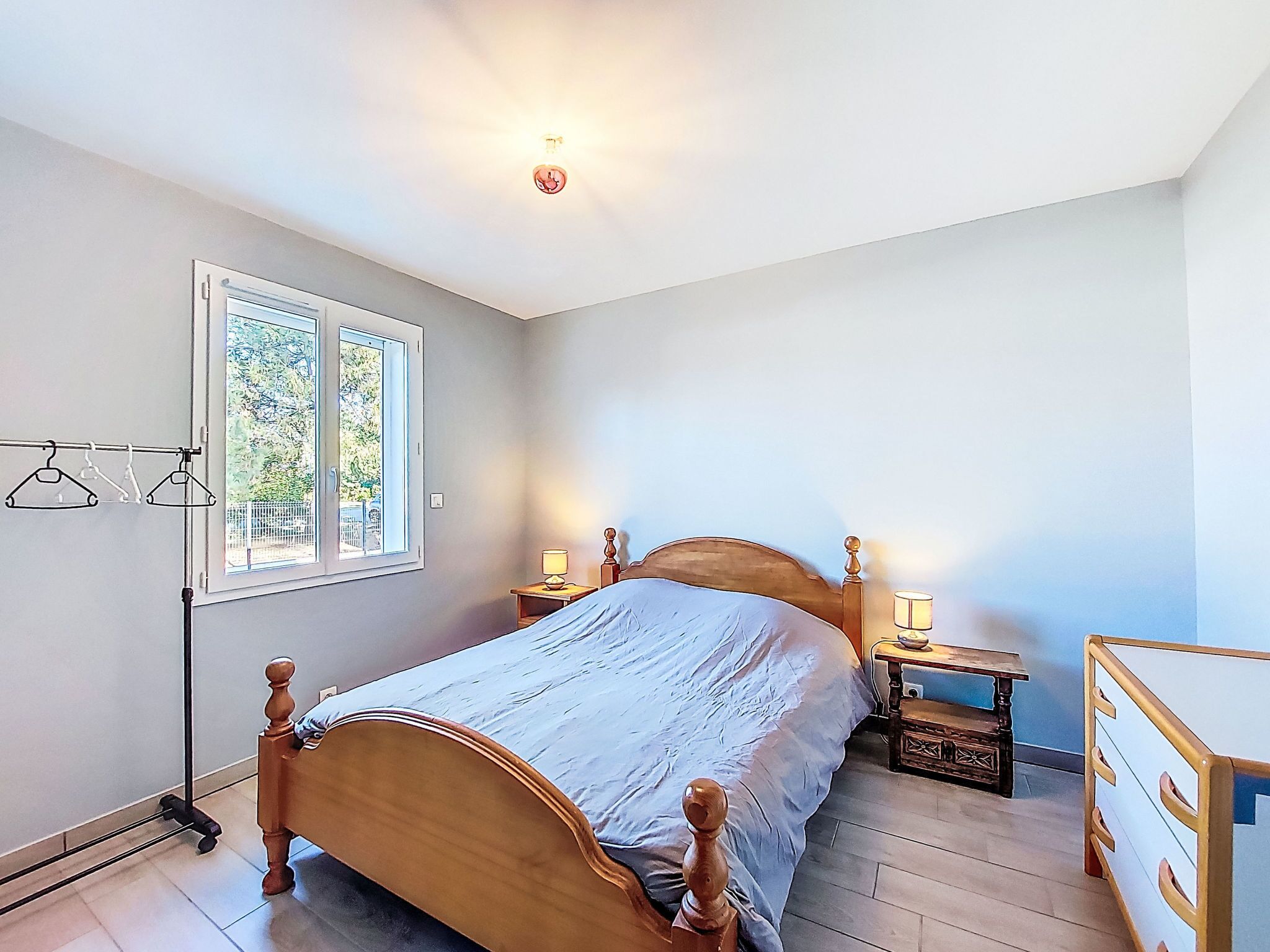 Quarto com cama de madeira, mesa de cabeceira com abajur, cômoda, radiador e janela com vista para vegetação.