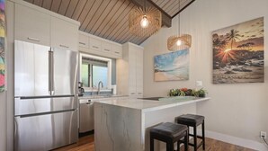 A modern kitchen with stainless steel appliances, a marble island, and a bar area with stools. - Aloha Condos, Keauhou Resort, Condo 118, Ocean View, AC (Kailua Kona)