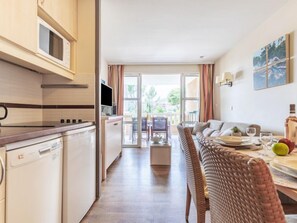 A modern kitchen with a dishwasher, microwave, and stove. There is a dining area with a table set for two, a sofa, and a view of the outdoors.