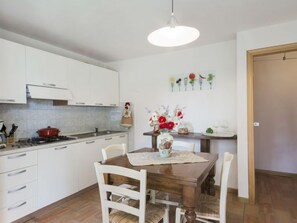 A kitchen with white cabinets, a wooden dining table with chairs, a hanging light fixture, and a vase with flowers on the wall.