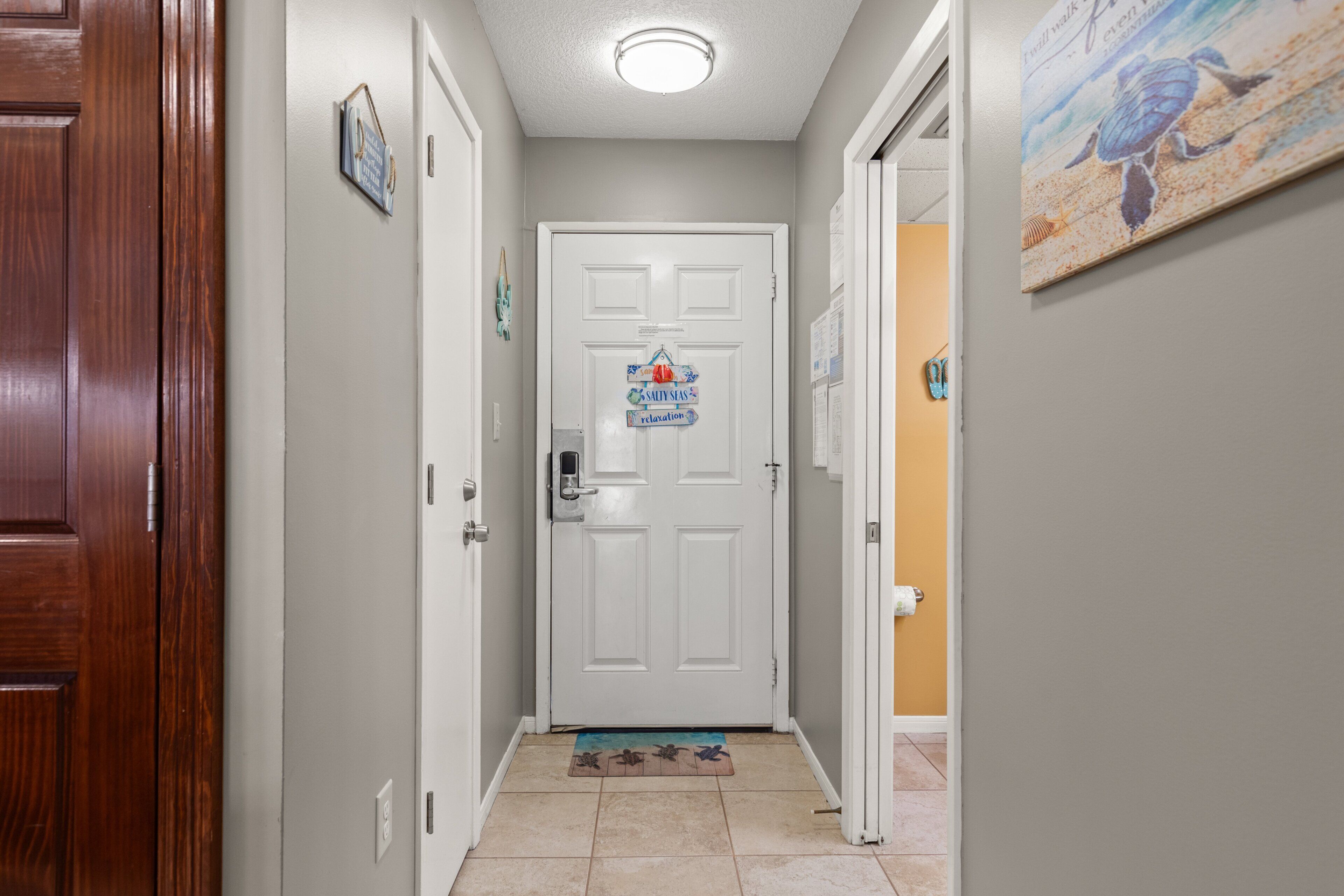 A hallway with a white door, a doormat, a framed sign, and a painting on the wall.