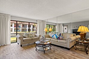Ein geräumiges Wohnzimmer mit zwei Sofas, einem Couchtisch und einem Fernseher. Große Fenster bieten Blick auf einen Innenhof.