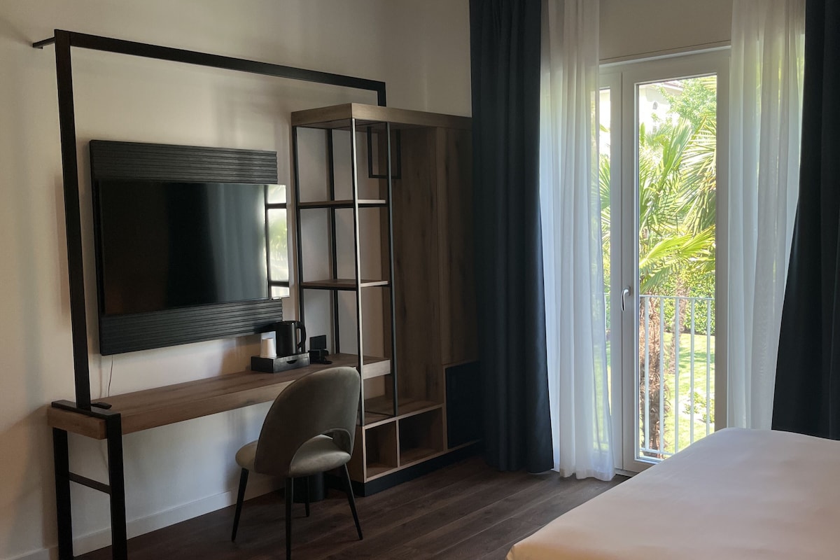 A modern hotel room with a large bed, a desk with a chair, a TV, and a sliding door to a balcony.
