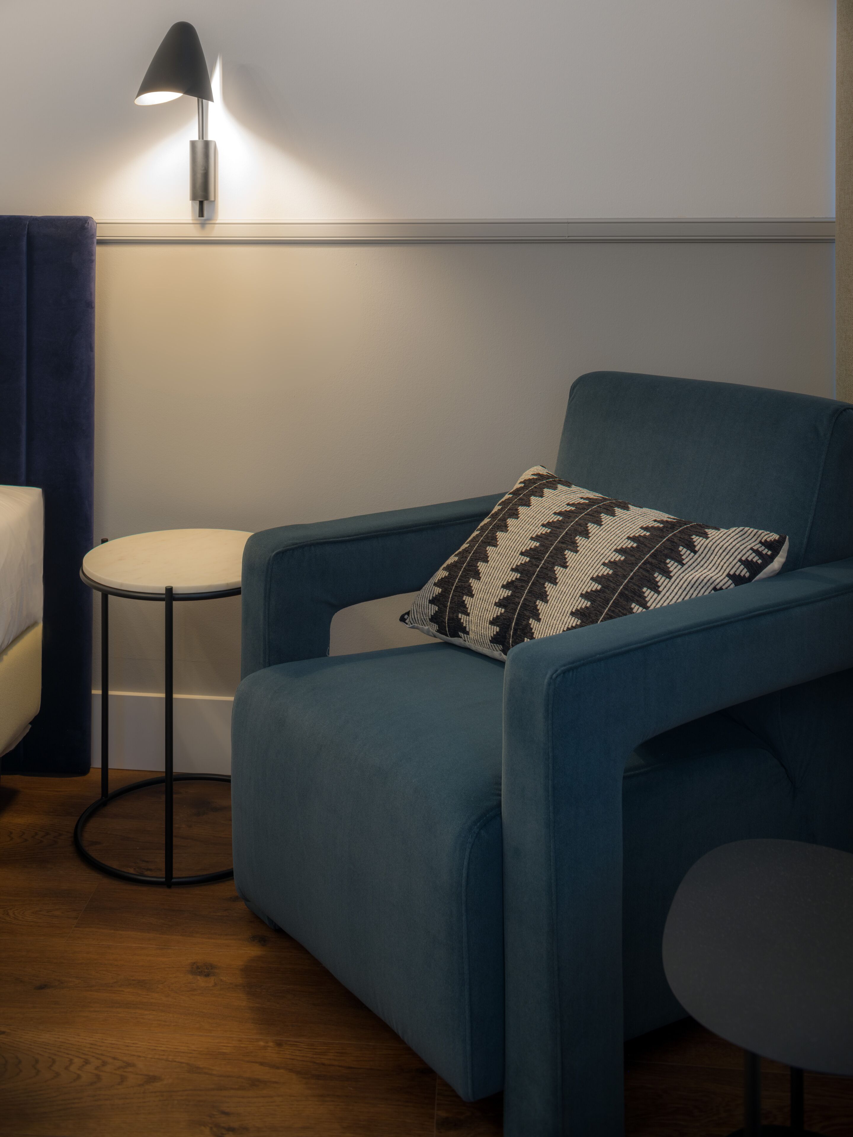 A blue armchair with a patterned pillow, a small round table, and a floor lamp in a room with wooden flooring.